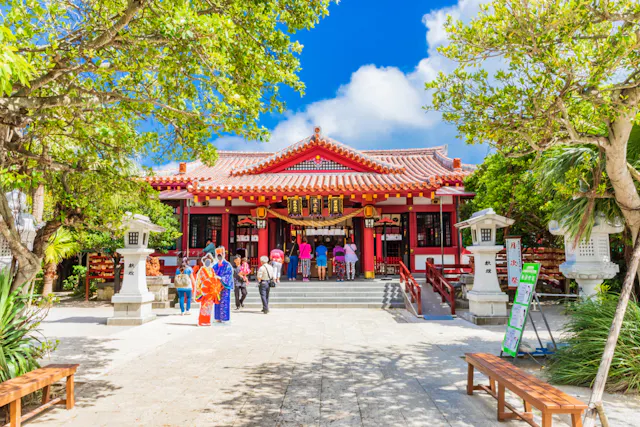 A group of people, some in traditional clothing, stand in front of a vibrant red and white Japanese shrine surrounded by trees under a blue sky with scattered clouds.