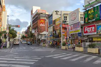 A busy urban street in Japan with colorful signs and billboards, including a McDonald's, shops, buses, pedestrians, and crosswalks under a partly cloudy sky.