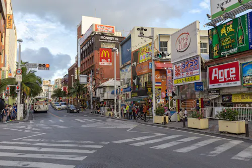 A busy urban street in Japan with colorful signs and billboards, including a McDonald's, shops, buses, pedestrians, and crosswalks under a partly cloudy sky.