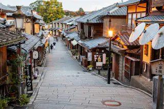 A narrow, stone-paved street lined with traditional wooden buildings and paper lanterns in a historic Japanese town, with a few people walking in the distance and warm streetlights illuminating the scene.