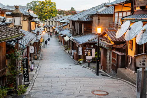 A narrow, stone-paved street lined with traditional wooden buildings and paper lanterns in a historic Japanese town, with a few people walking in the distance and warm streetlights illuminating the scene.