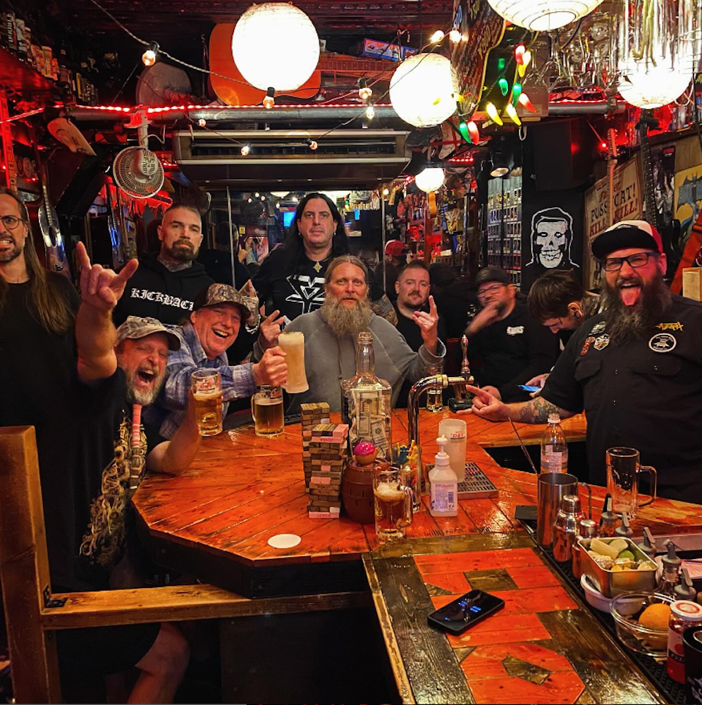 A vibrant bar scene with a diverse group of cheerful people sitting around a wooden hexagonal bar table, raising their drinks and making rock-on hand gestures. The bar is warmly lit with colorful decorations, bottles, and memorabilia adorning the walls.