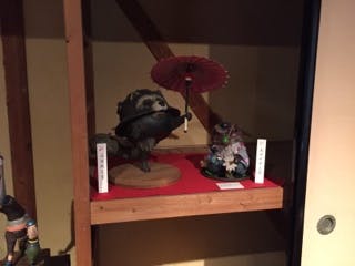 A figurine of a raccoon dog holding a red paper umbrella is displayed on a red shelf, next to another detailed figurine with Japanese labels in a wooden display case.