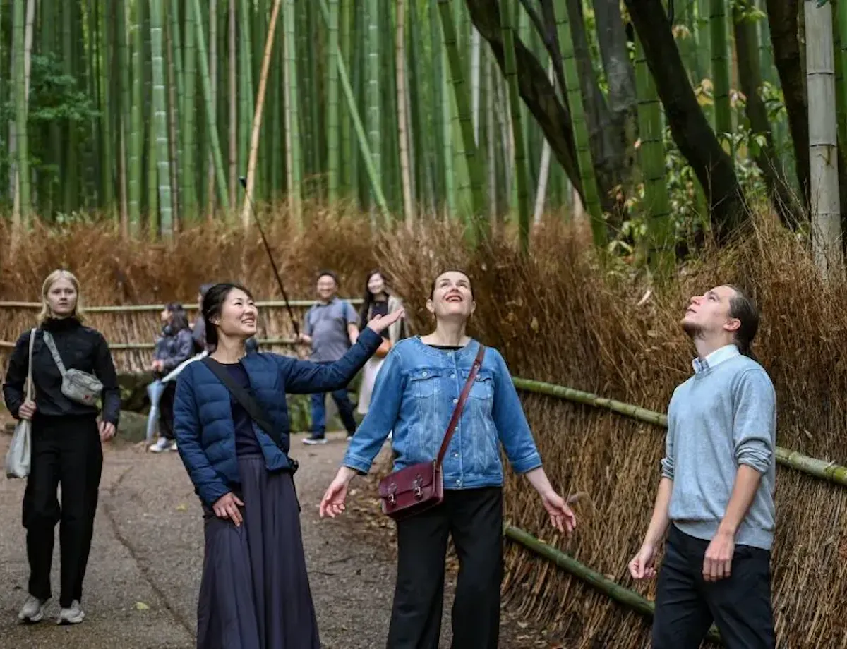 Arashiyama Bamboo Grove A group of people walks along a path through a dense bamboo forest, some looking up in awe at the tall bamboo stalks surrounding them. The atmosphere appears peaceful and serene.