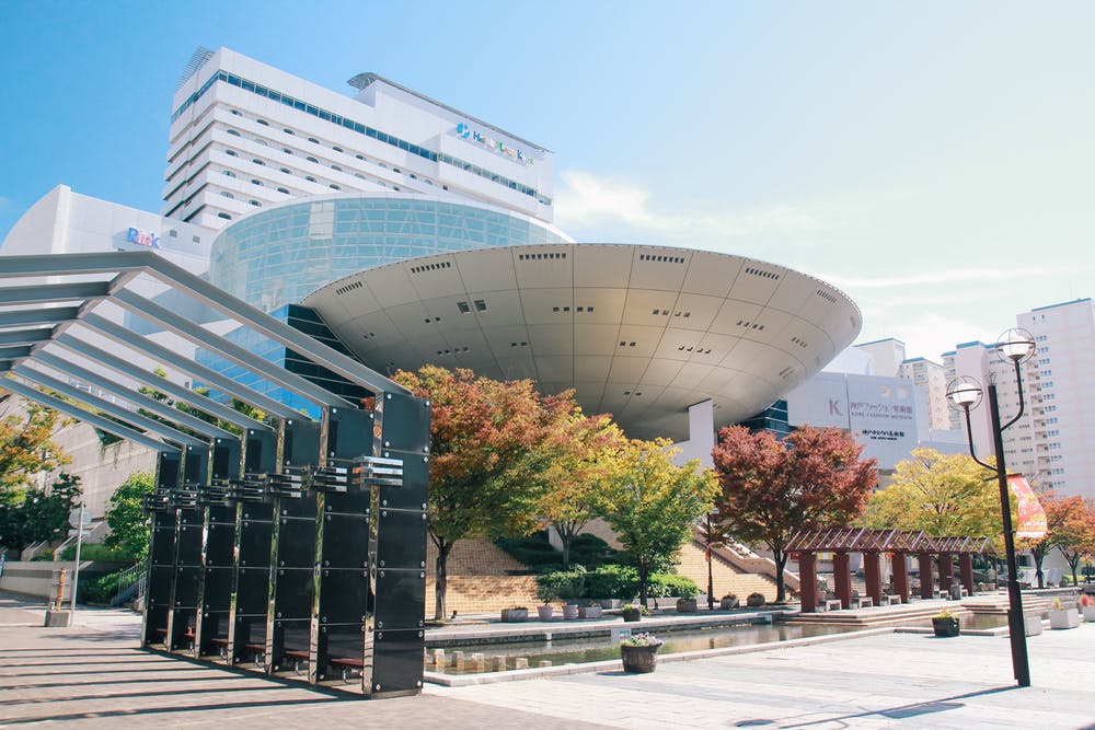 A modern urban plaza featuring a futuristic, circular building with glass and metal elements, surrounded by trees with autumn foliage and nearby tall buildings under a clear blue sky.