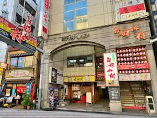 Street view of a commercial building in Japan with various colorful signs in Japanese, storefronts, and entrances to restaurants at ground level. The area appears lively and urban.