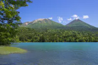 A clear blue lake bordered by green trees with two forested mountains in the background under a bright blue sky with a few clouds.