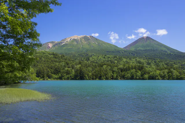 A clear blue lake bordered by green trees with two forested mountains in the background under a bright blue sky with a few clouds.