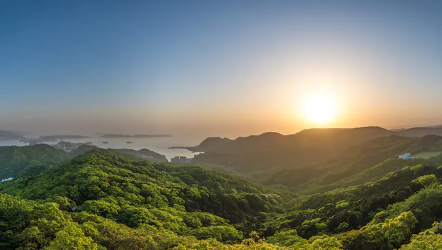 The sun rises over green, tree-covered hills with a clear sky, casting warm light on the landscape. In the distance, water and small islands are visible, creating a peaceful, scenic view.