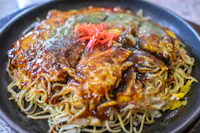 A close-up of a Japanese okonomiyaki pancake topped with sauce, noodles, and garnished with red pickled ginger, served on a black plate.