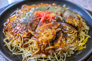 A close-up of a Japanese okonomiyaki pancake topped with sauce, noodles, and garnished with red pickled ginger, served on a black plate.
