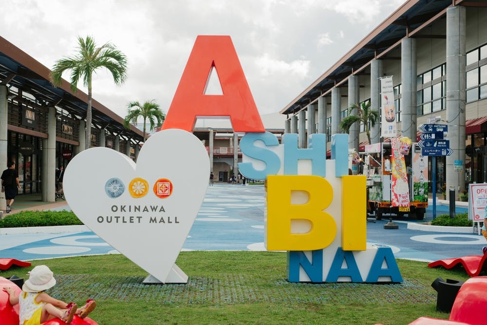 Colorful "ASHIBINAA" sign and white heart display for Okinawa Outlet Mall stand on grass, surrounded by palm trees and modern mall buildings under a cloudy sky. A child in a hat sits in the foreground.