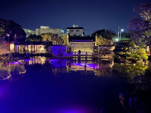 A tranquil garden at night with traditional Asian architecture, illuminated buildings, and vibrant purple reflections on a calm pond, surrounded by trees and city structures in the background.