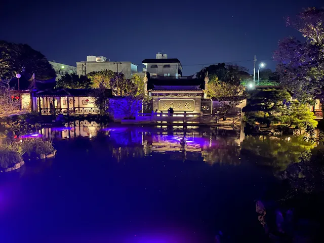 A tranquil garden at night with traditional Asian architecture, illuminated buildings, and vibrant purple reflections on a calm pond, surrounded by trees and city structures in the background.