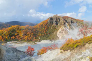 A steaming hot spring surrounded by colorful autumn trees and hills under a partly cloudy sky, with vibrant red, orange, and yellow foliage blanketing the landscape.