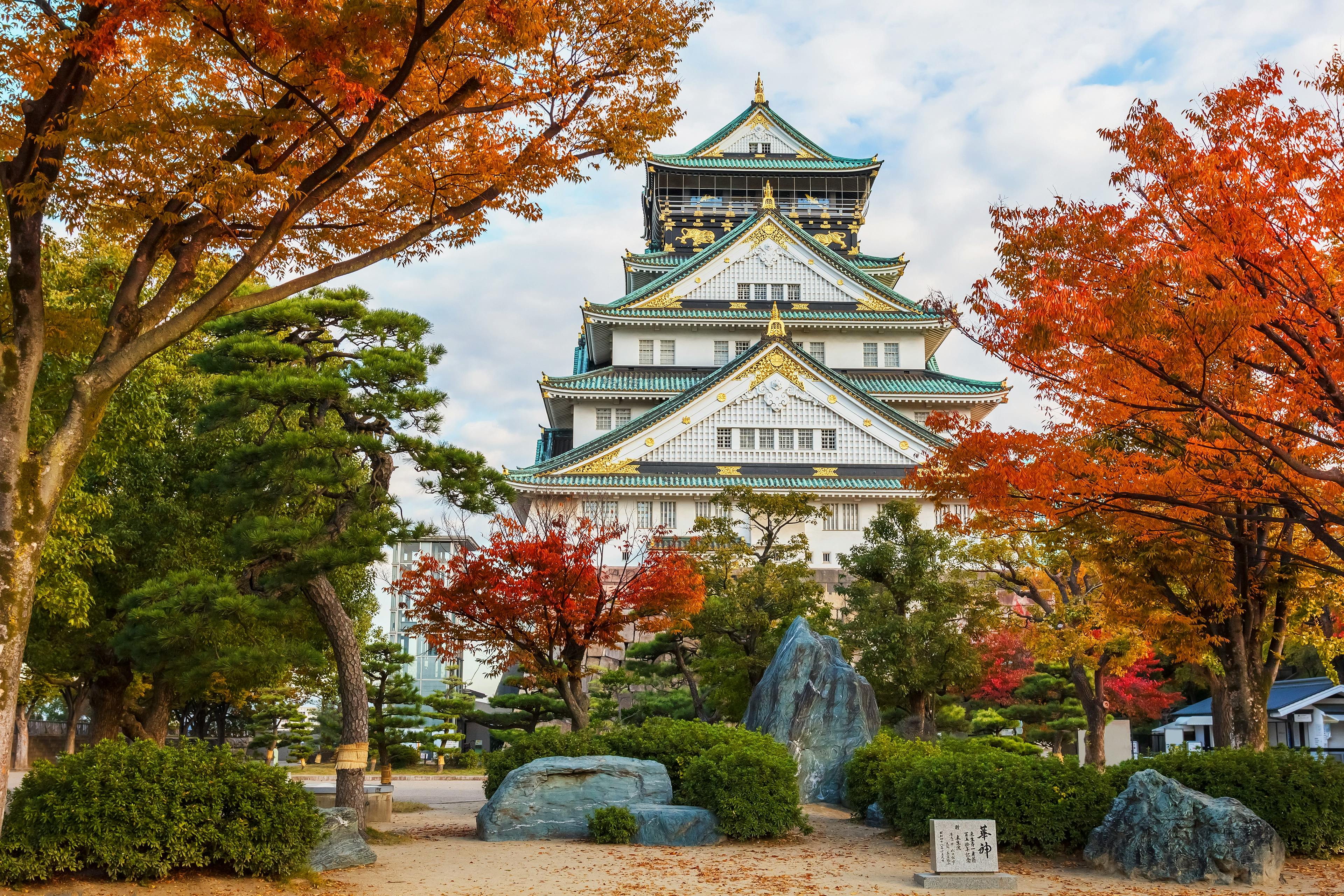 Osaka Castle in Japan surrounded by colorful autumn trees, with red and orange leaves, blue sky with clouds, and rocks and bushes in the foreground.