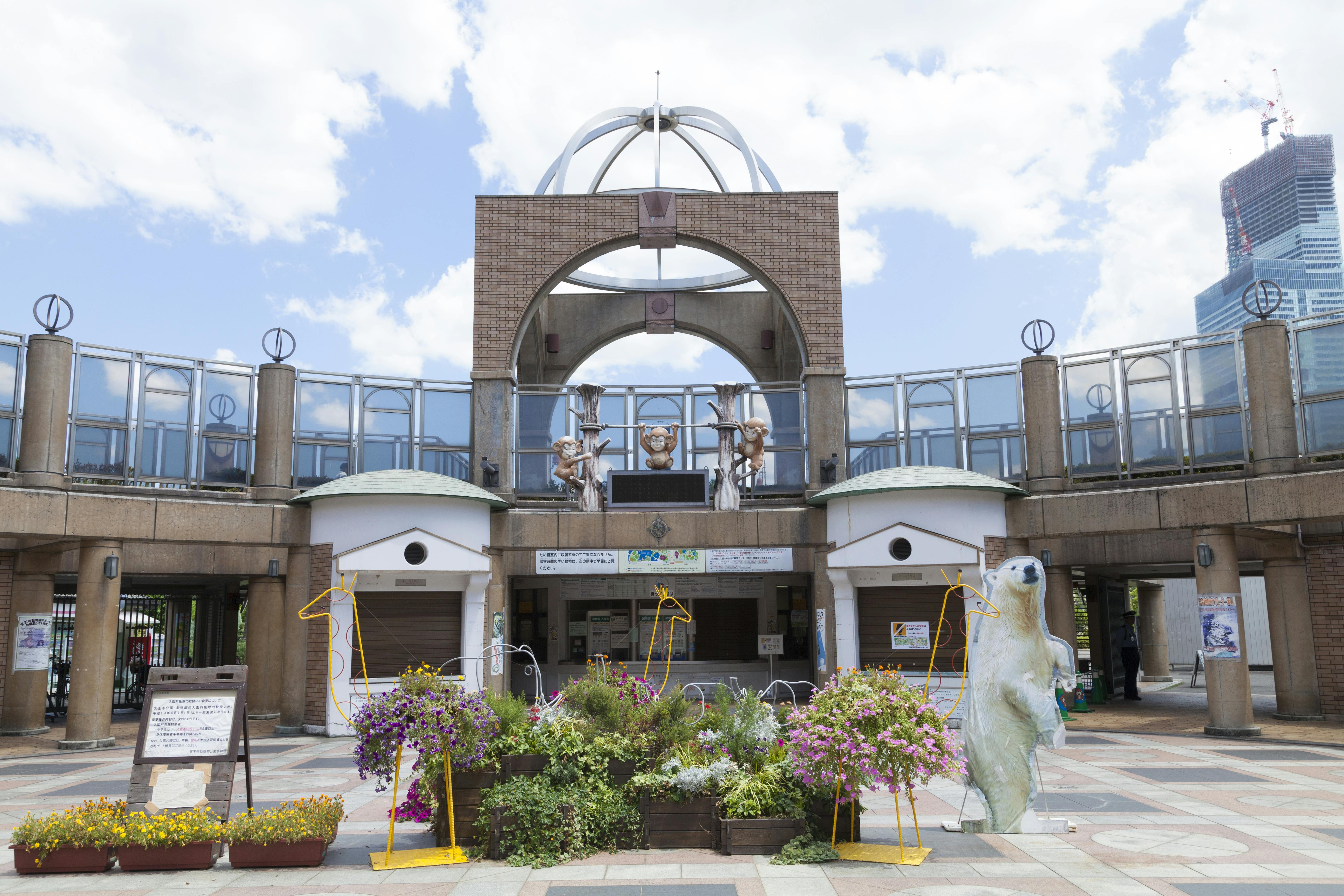 Public square with a circular plaza, featuring flower arrangements and a large polar bear sculpture in the center. The arched structure behind has decorative elements. A tall building is visible in the background under a partly cloudy sky.