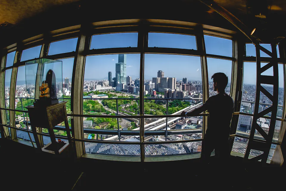 A person stands in a high-rise building, looking out through large windows at a cityscape. The view includes various skyscrapers, a green park area, and a clear blue sky. A display case is visible to the left.
