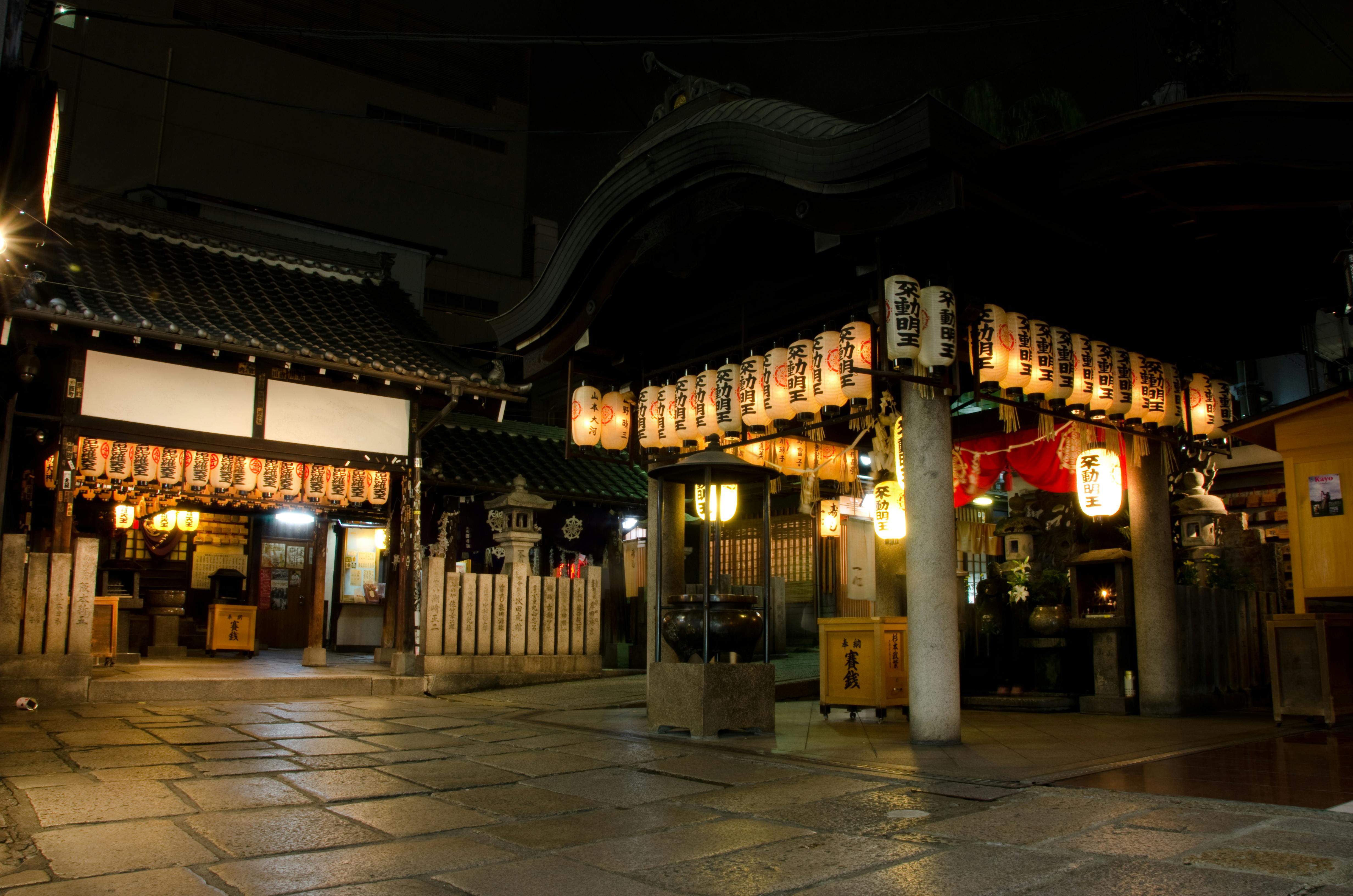 Nighttime view of a traditional Japanese shrine, illuminated by numerous hanging lanterns with Japanese characters. The stone courtyard and wooden structures create a serene atmosphere.