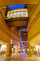 Modern indoor plaza with curved stone walls, glass skywalk above, and colorful lights illuminating the space. Shops line the walkway, and people move through the vibrant area in the evening.