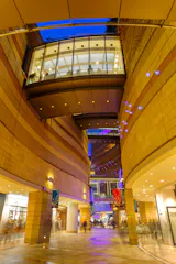 Modern indoor plaza with curved stone walls, glass skywalk above, and colorful lights illuminating the space. Shops line the walkway, and people move through the vibrant area in the evening.
