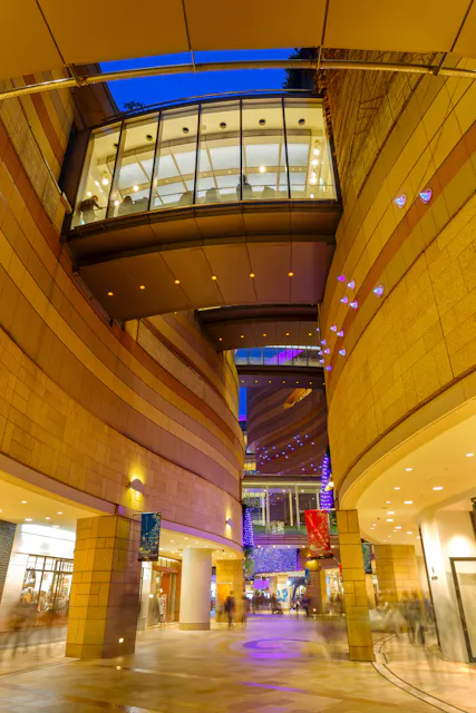 Modern indoor plaza with curved stone walls, glass skywalk above, and colorful lights illuminating the space. Shops line the walkway, and people move through the vibrant area in the evening.