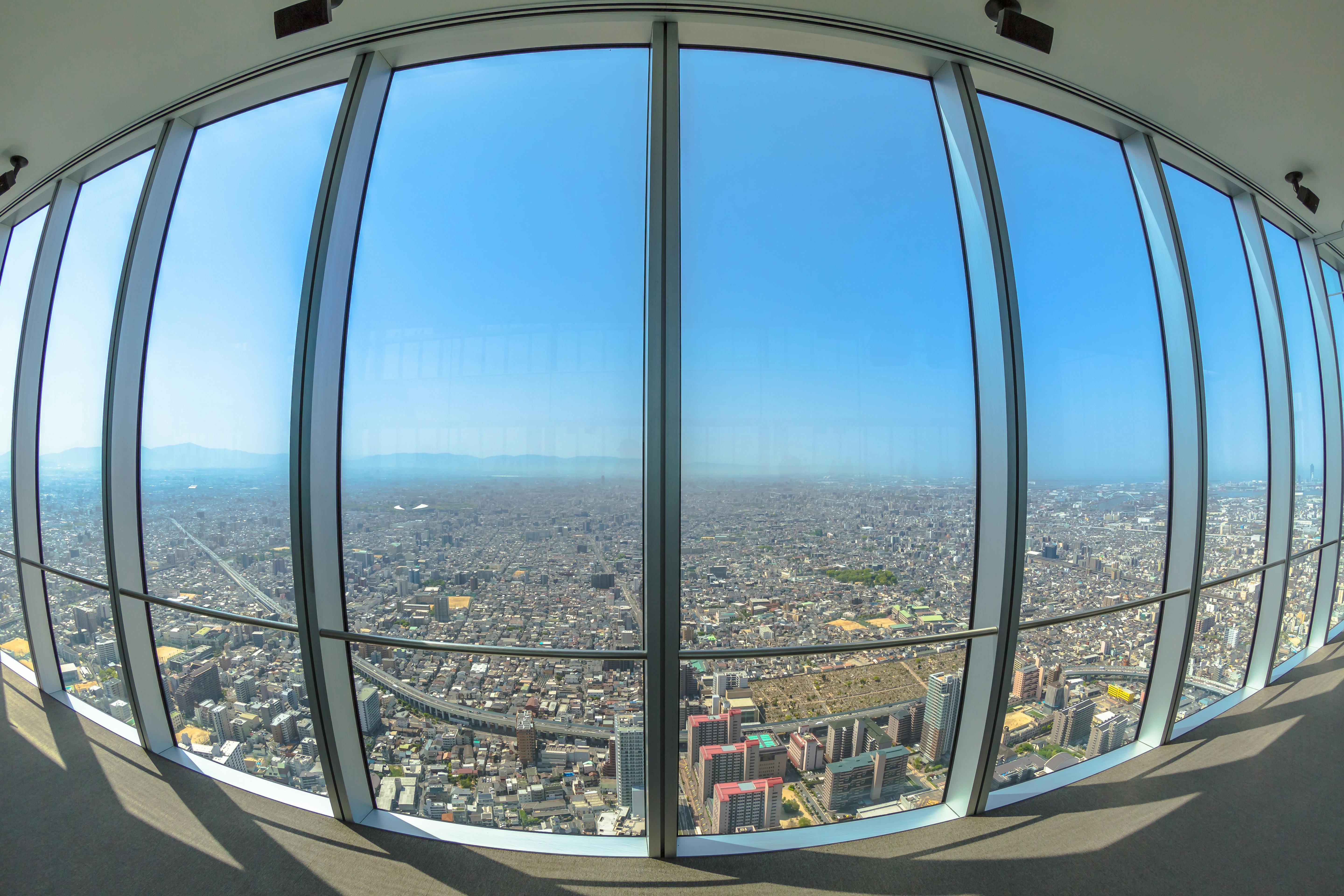 Wide cityscape view through large, curved floor-to-ceiling windows of a tall observation deck, overlooking a sprawling urban area under a clear blue sky.
