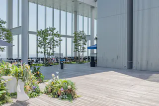 A modern outdoor terrace with wooden flooring, potted plants, tall glass windows, and a seating area shaded by blue canopies. Tall columns and vertical metal panels add to the contemporary design.