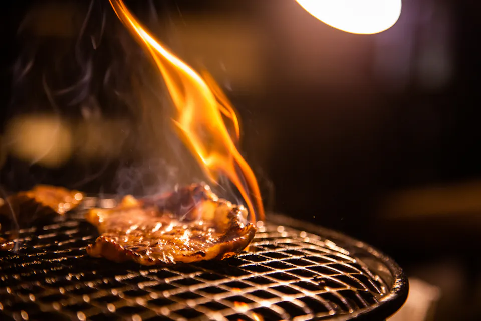 オープンフレームで肉がグリルされているクローズアップ。一片の肉が際立っており、その上に炎が立ち上り、グリルの周りには目に見える煙があります。光源がシーンの一部を照らし、グリルプロセスに重点を置いています。