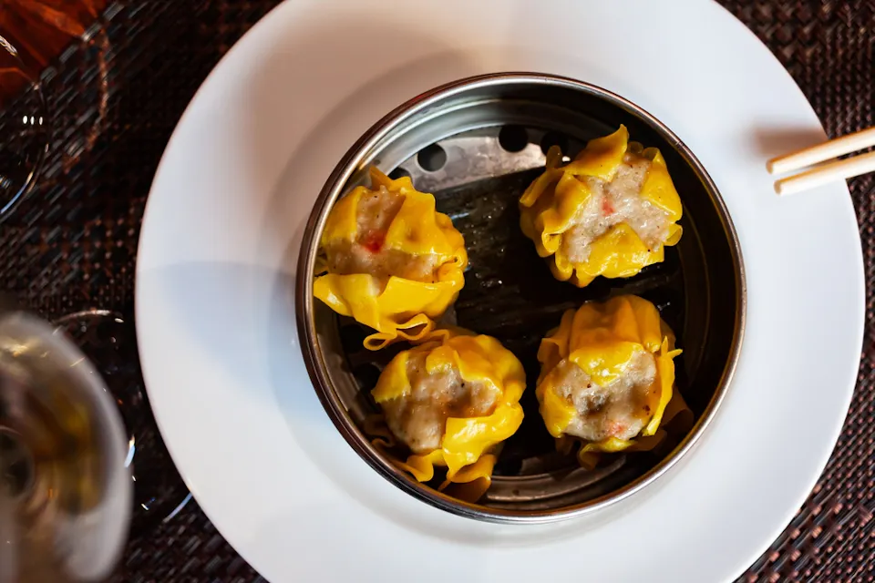 クリーミーな具が入った4つの蒸したての黄色い餃子の上からの視点。蒸し器は白い皿の上に置かれ、端には箸があり、左側には部分的に見えるグラスがある。