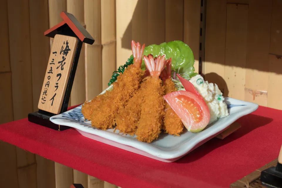 レタス、ポテトサラダ、トマトのくさびを添えた衣をつけて揚げたエビの皿が赤い表面に展示されています。 plateの横に日本語の書かれた木のサインがあります。背景は竹の壁です。