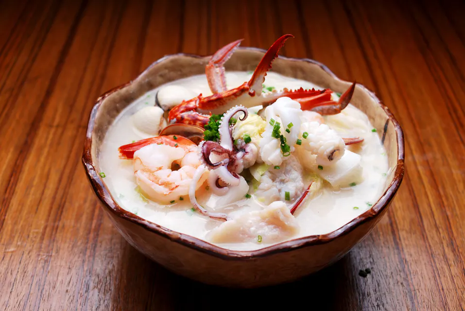 木のテーブルの上にクリーミーなシーフードスープが盛られ、カニの爪、エビ、イカ、魚の塊が刻んだねぎで飾られています。濃厚なスープは白く、少しとろみがあり、ボリューム感のある見た目を作り出しています。
