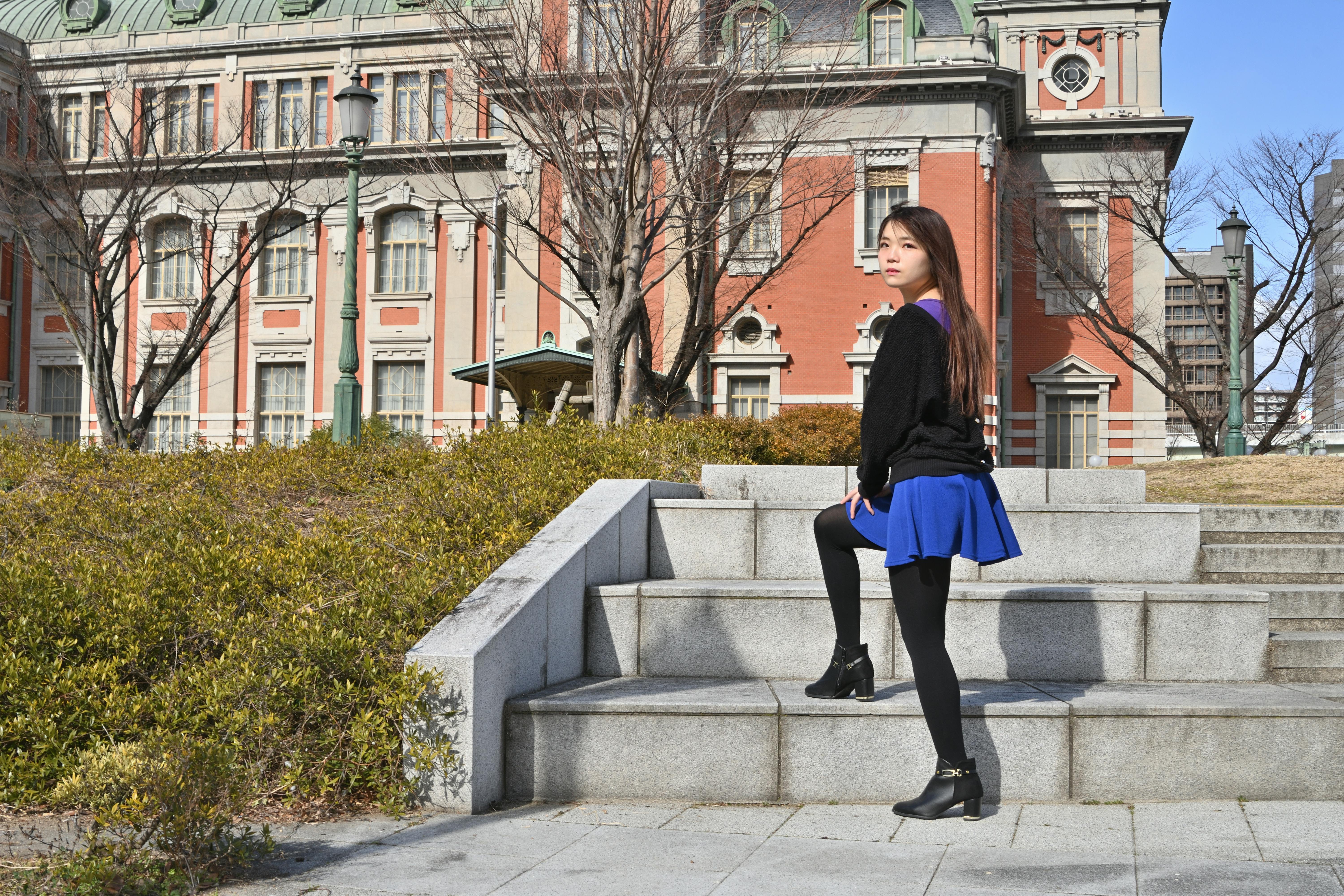 A young woman in a blue skirt and black sweater stands on stone steps outdoors, posing with one foot raised. Behind her is a large, historic brick building with green roofs and leafless trees.