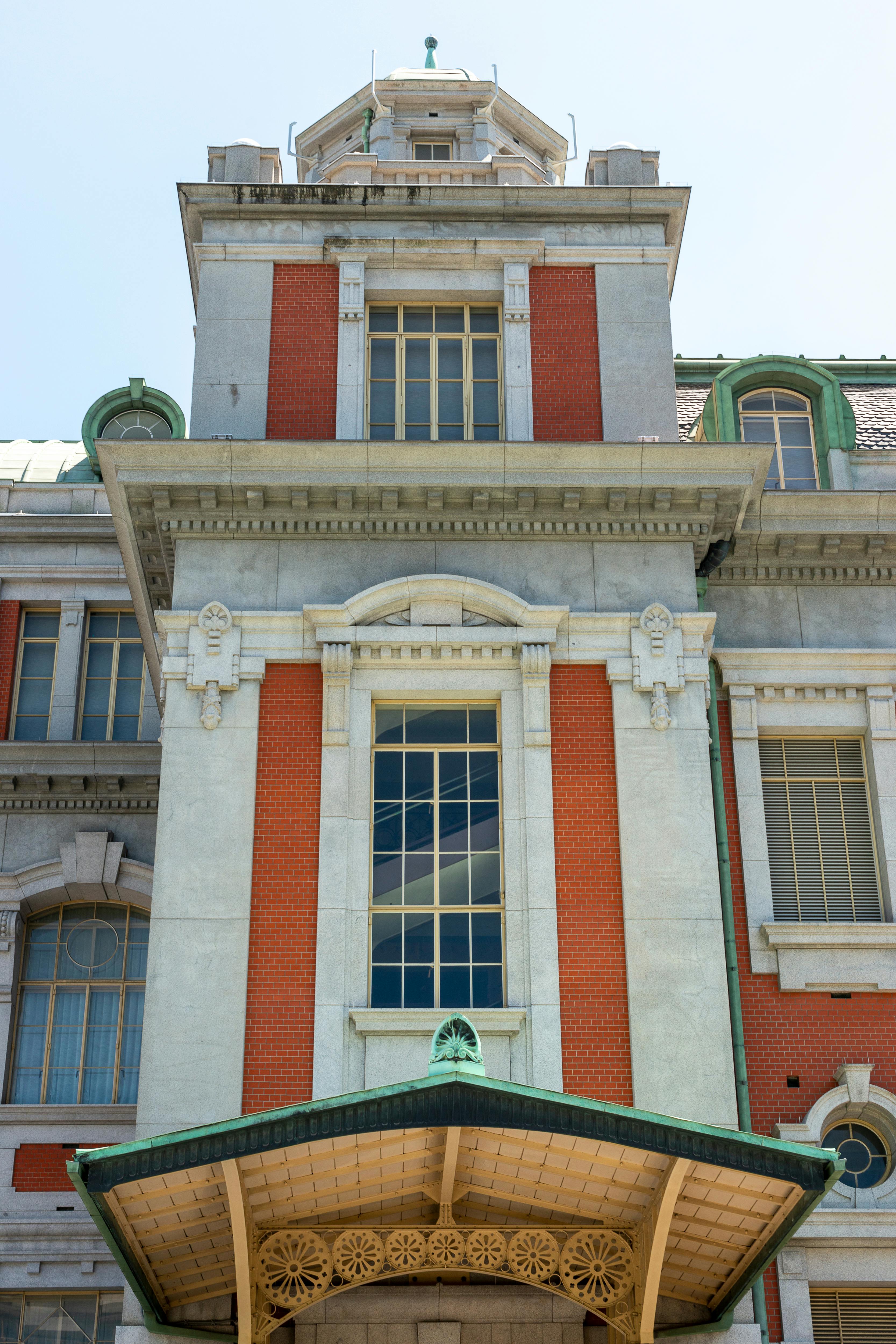 A historic building facade with red brick and white stone, featuring tall windows, decorative molding, and a green roof with an ornate canopy above the entrance.