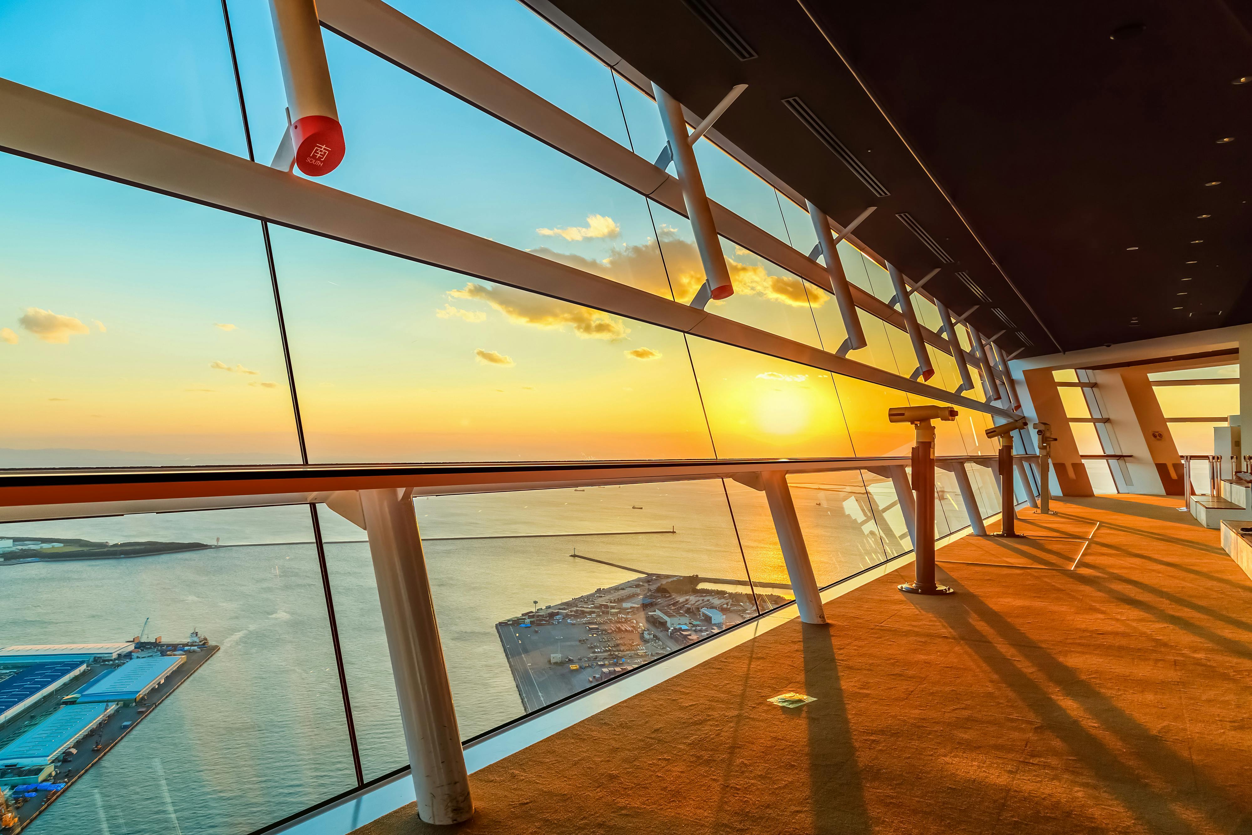 A modern observation deck with large glass windows overlooks a harbor at sunset, casting warm golden light across the floor. A telescope stands near the windows, and the sun is low on the horizon above the calm water.