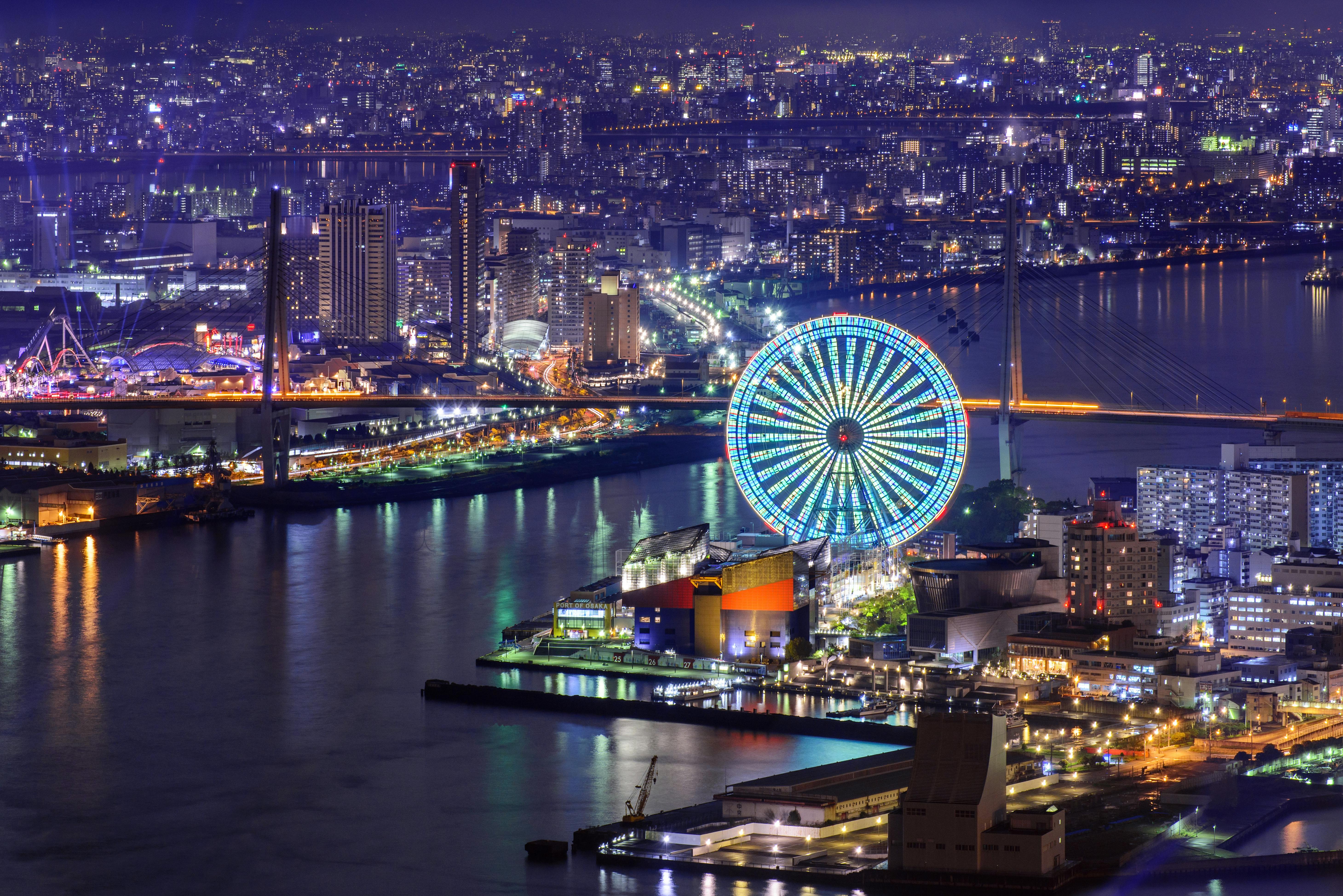 A vibrant cityscape at night featuring a large, brightly lit Ferris wheel by the waterfront, surrounded by illuminated buildings, bridges, and reflections on the water.
