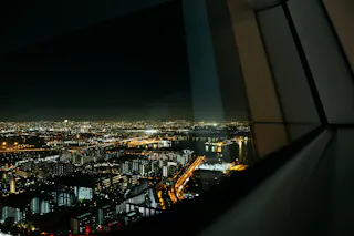 A nighttime cityscape with illuminated buildings and streets, seen through a window with reflections on the glass, overlooking a waterfront area and distant lights.
