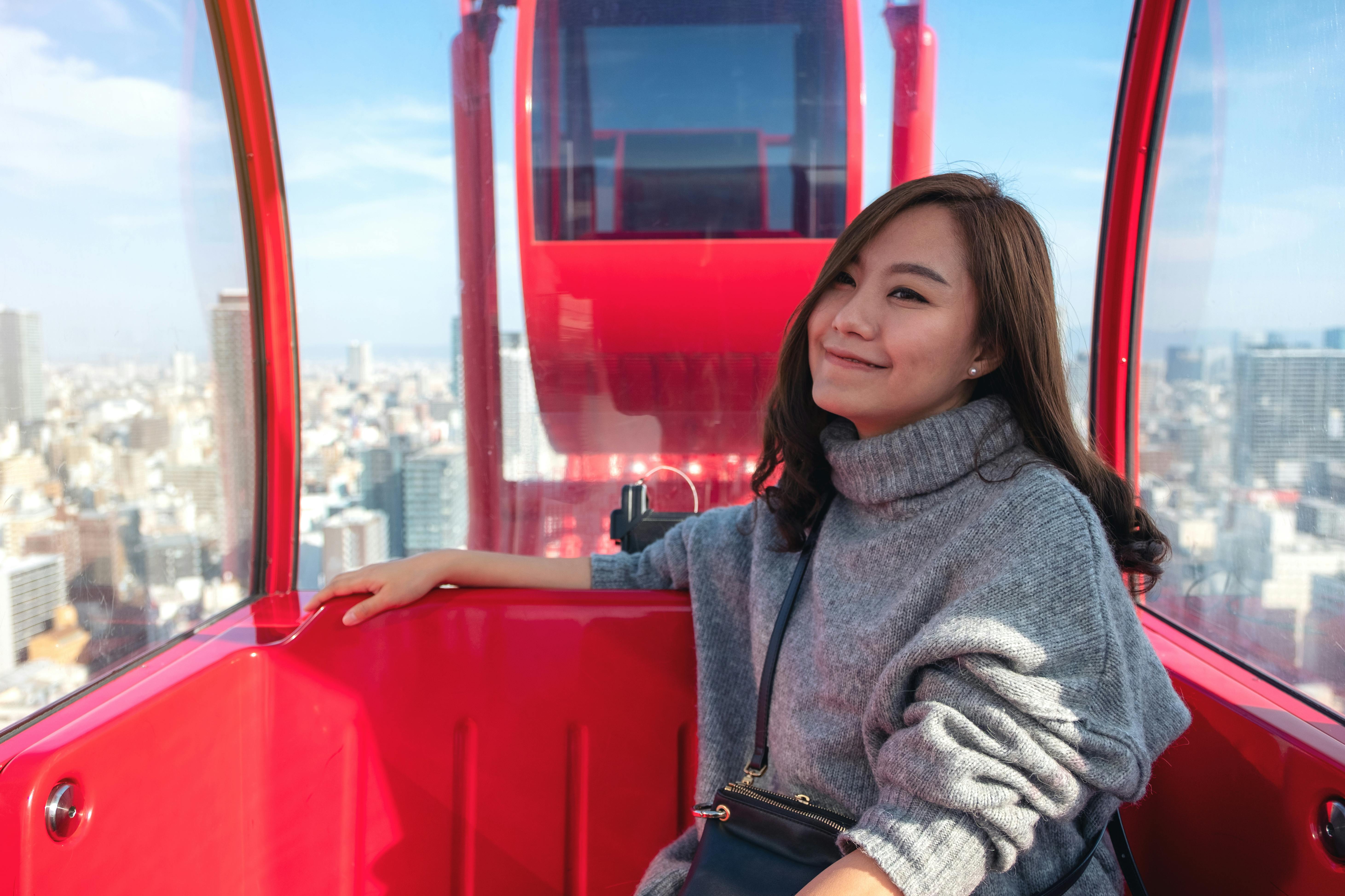 A woman wearing a gray sweater smiles while sitting inside a bright red Ferris wheel cabin, with a cityscape and blue sky visible through the window behind her.
