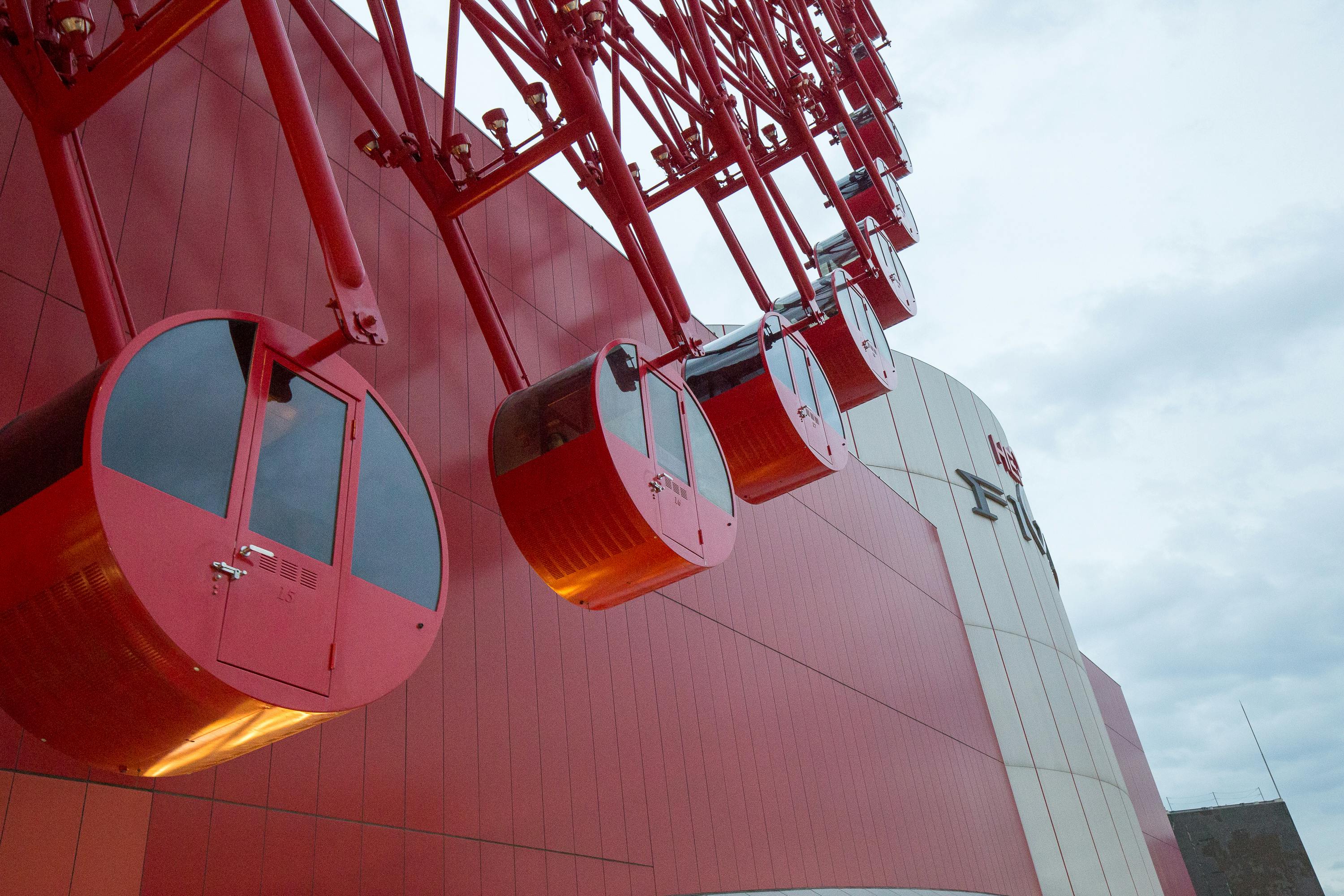Red Ferris wheel cabins partially elevated against a modern red and white building with curved architecture, set against a cloudy sky. The angle is from below, highlighting the structure and glass panels.