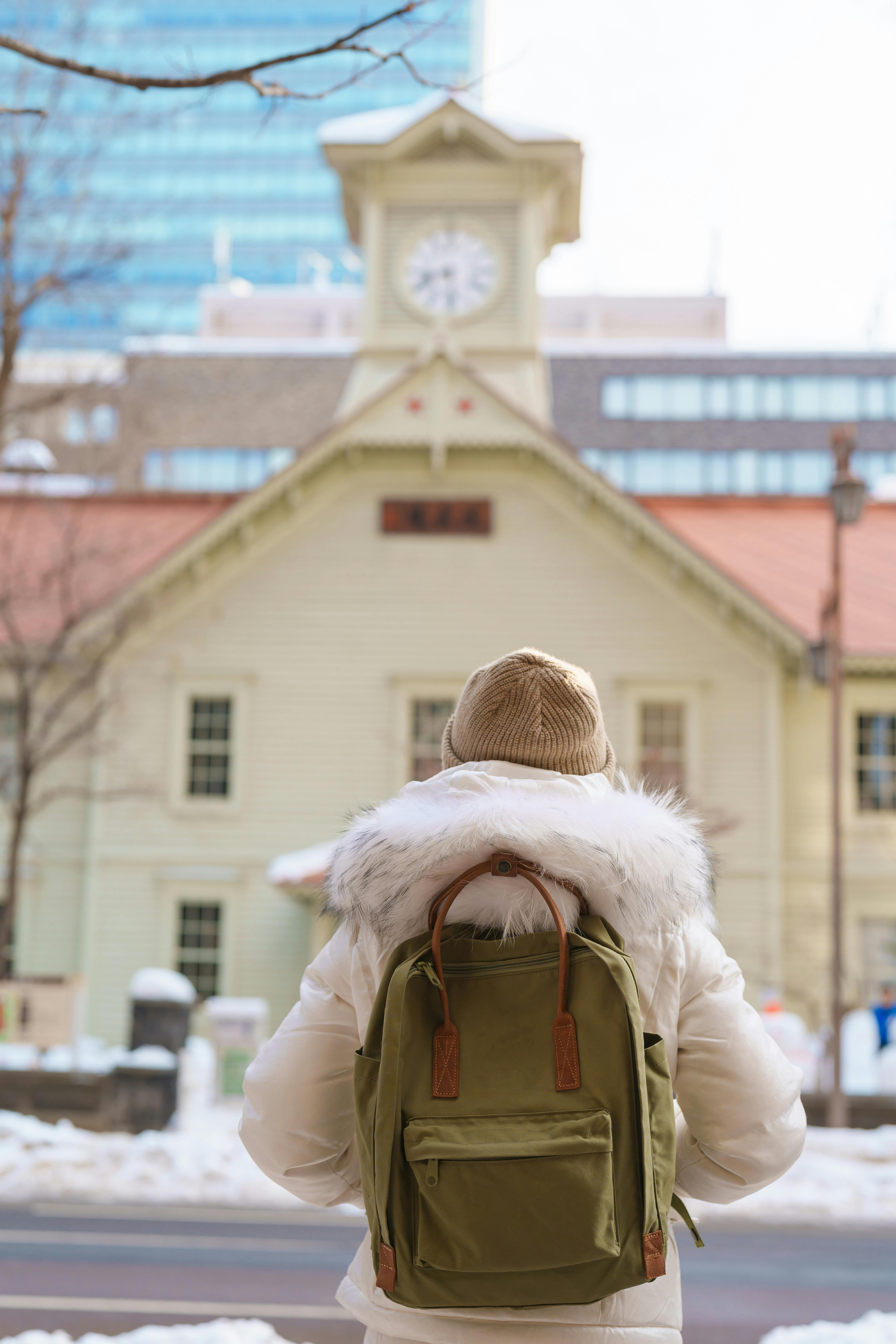 A person wearing a white coat, beige hat, and green backpack stands facing a historic clock building on a snowy day in the city.