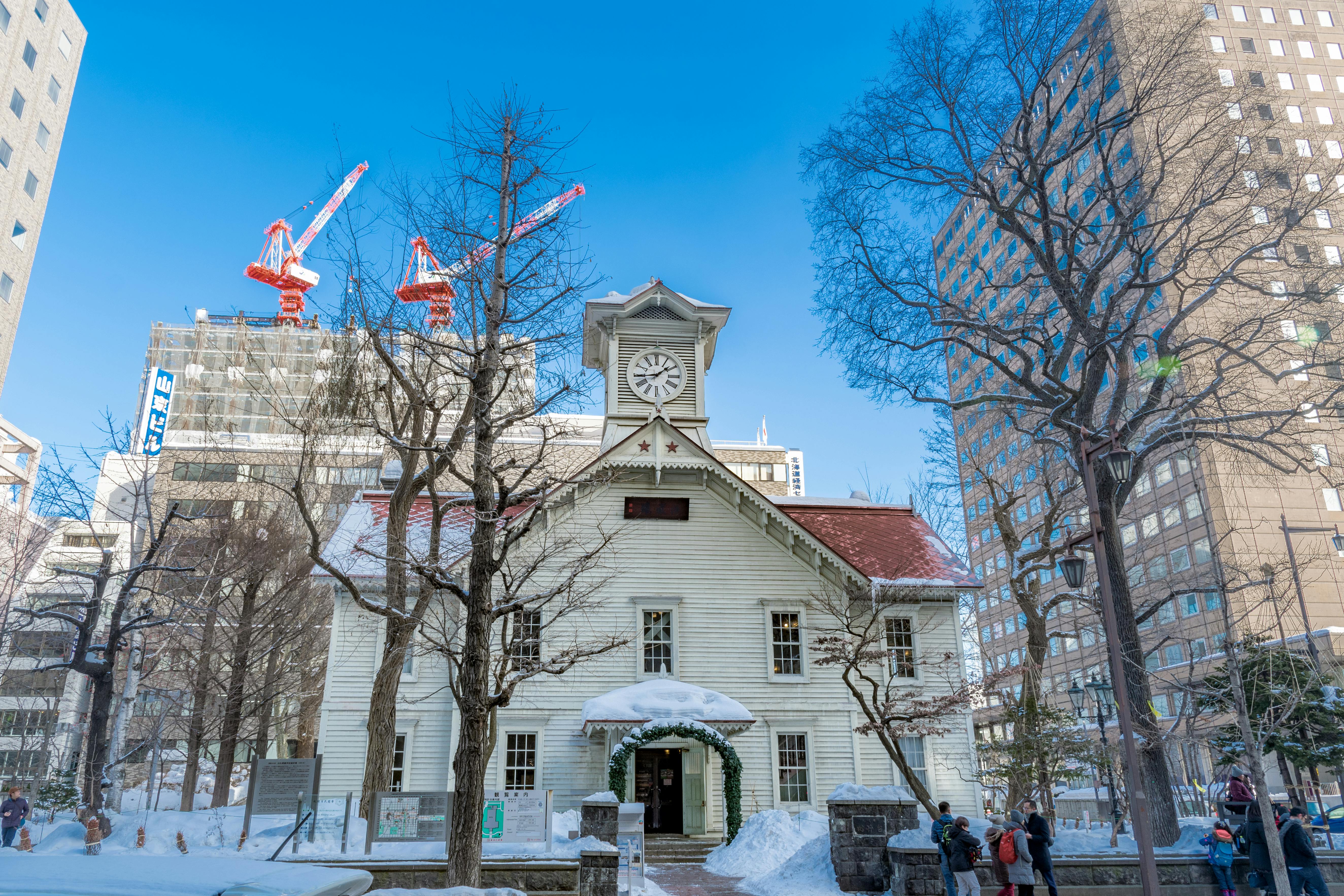 A historic white wooden clock tower building stands amid snow and leafless trees, surrounded by modern high-rise buildings and construction cranes under a clear blue sky in an urban setting.