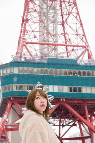 A woman in a white coat stands in front of a red steel tower, possibly Tokyo Tower, with snow falling and snow-covered structures visible in the background.