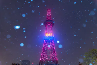 A tall tower illuminated with pink lights displays the time "10:41" on a digital clock, as snow falls heavily at night. The sky is dark, and the glowing lights contrast with the scattered snowflakes.