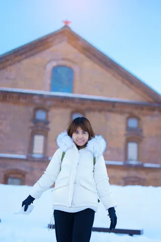 A young woman wearing a white winter coat with a fur-lined hood and black gloves stands smiling in front of a snow-covered brick building on a bright winter day.