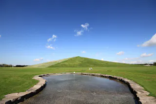 A small circular pond with a stone border sits in the foreground of a wide green field, with a large grassy hill rising under a clear blue sky with scattered clouds.