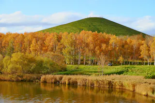 A grassy hill with people standing on top, surrounded by autumn trees with golden leaves, a pond in the foreground, and a car parked near the trees under a blue sky with scattered clouds.