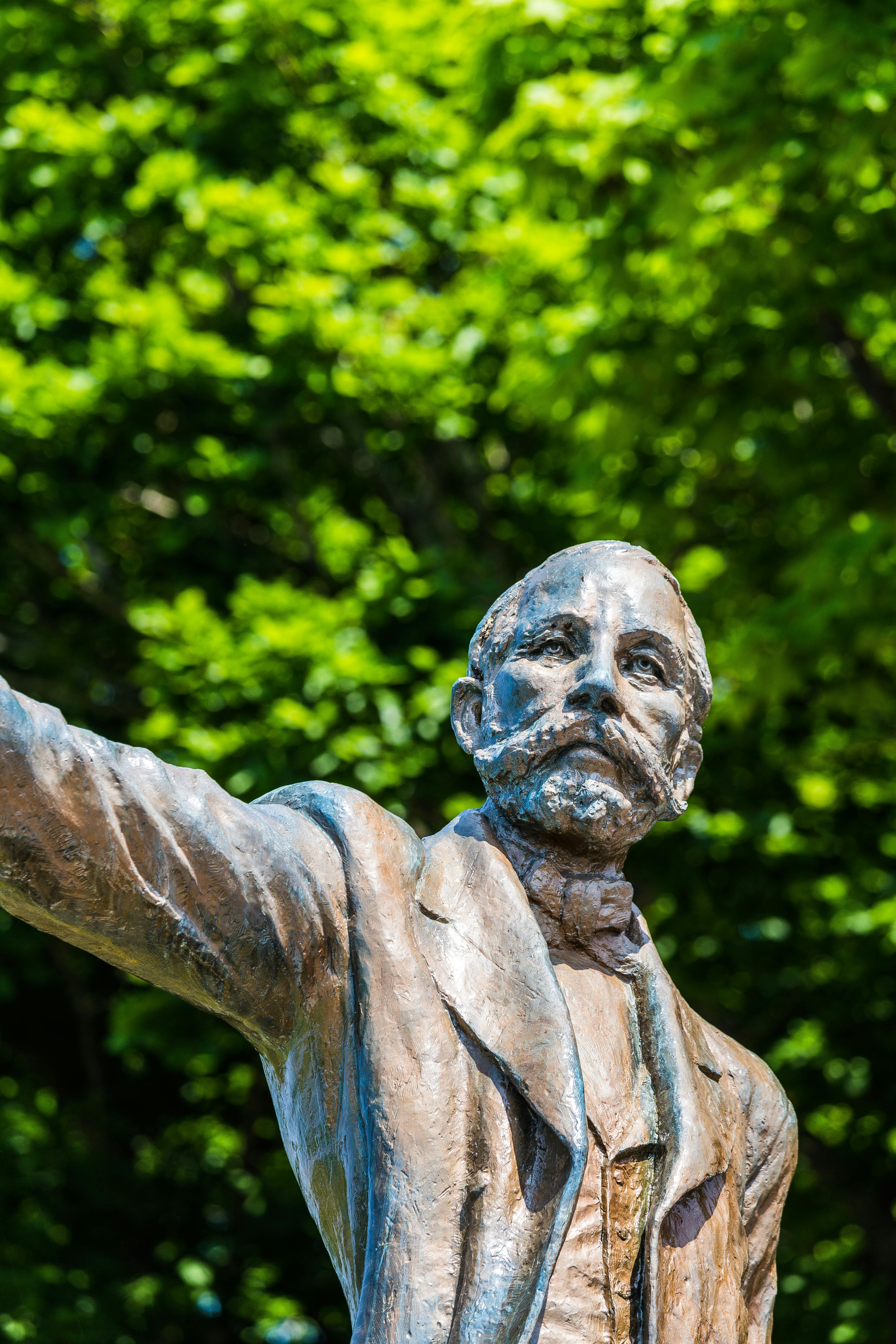 A bronze statue of a bearded man in a suit with one arm extended, set against a background of lush green trees and bright sunlight.