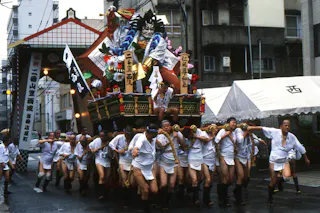 A group of people in traditional Japanese clothing pull a decorated festival float with colorful figures through a city street during a festival. Some participants push from behind, and buildings are visible in the background.