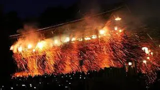 A nighttime festival scene shows bright orange sparks and fire raining down from a wooden structure, illuminating a crowd of people watching below.
