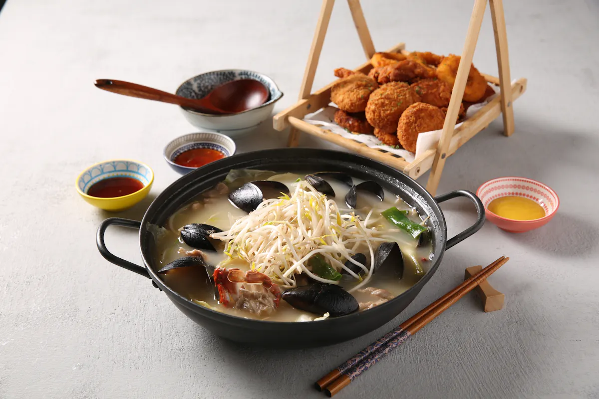 A steaming hot pot filled with seafood, vegetables, and bean sprouts is placed on a table alongside small bowls of dipping sauces and condiments. In the background, a wooden rack holds a serving of crispy fried snacks. Chopsticks rest on the table nearby.